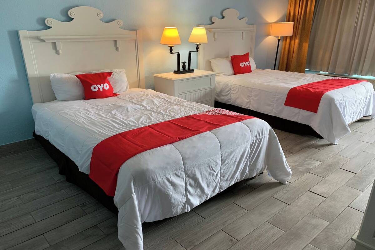 Deux lits avec une literie blanche et des touches de rouge, une table de chevet avec des lampes, et une vue sur l’extérieur par une fenêtre ornée de rideaux.