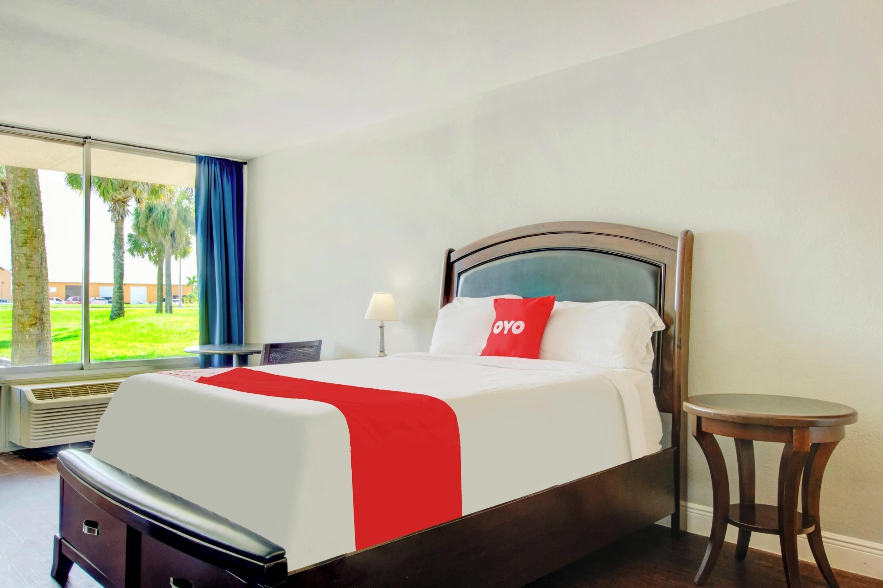 A hotel room with a bed, a small table, a lamp, and a view of palm trees through a window.