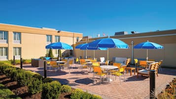 Een terras buiten met gele en oranje stoelen en tafels, blauwe parasols en een gebouw met ramen op de achtergrond.