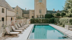 Une piscine avec des chaises longues, un bâtiment en pierre doté de fenêtres en arc de cercle et un ciel dégagé.