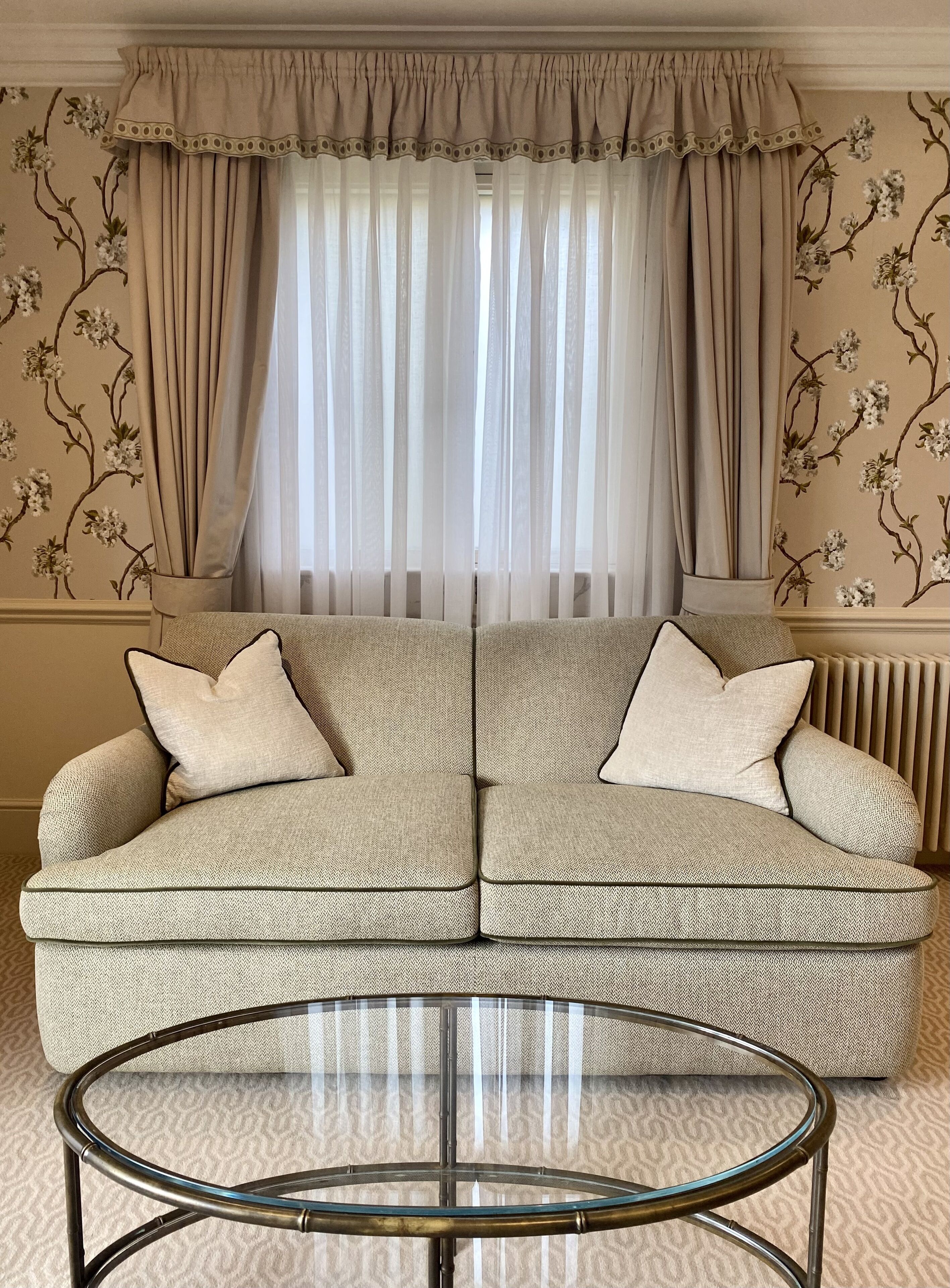Un salon avec un canapé beige, deux coussins, une table basse en verre et une fenêtre ornée de rideaux légers.