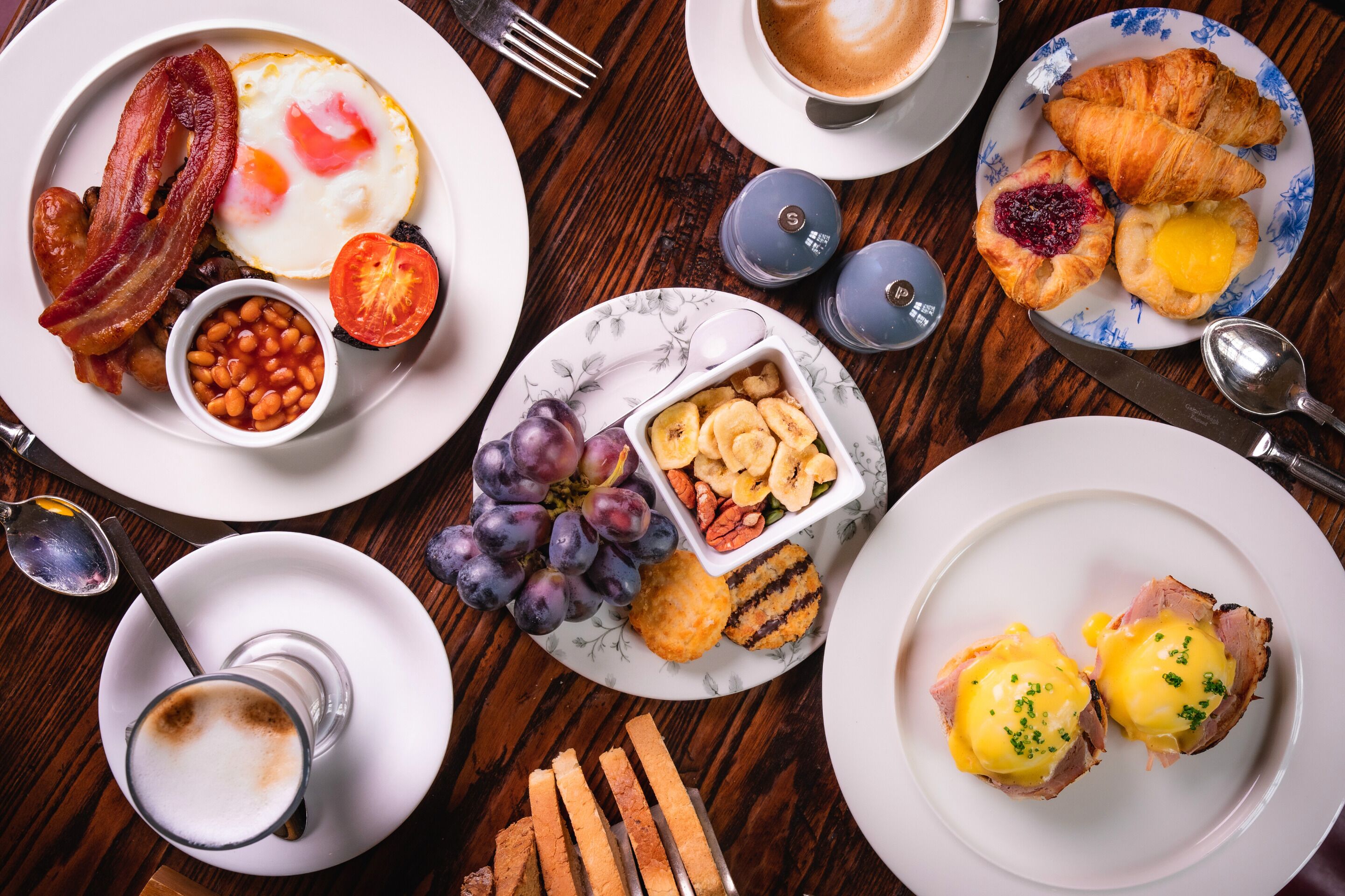 A breakfast spread with bacon, eggs, toast, and pastries on a wooden table.