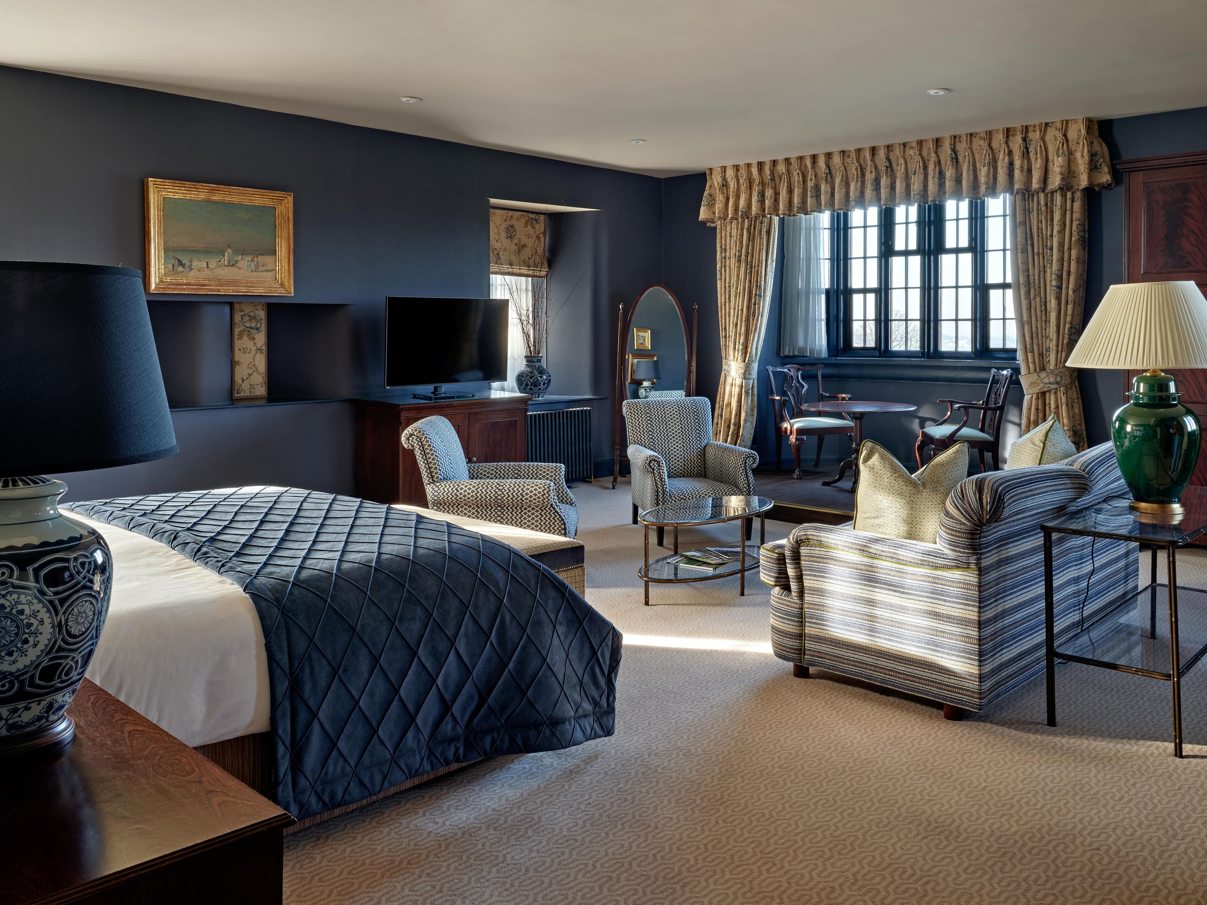 A hotel room with a large bed, two armchairs, a sofa, a small table, a TV, and a window with curtains.