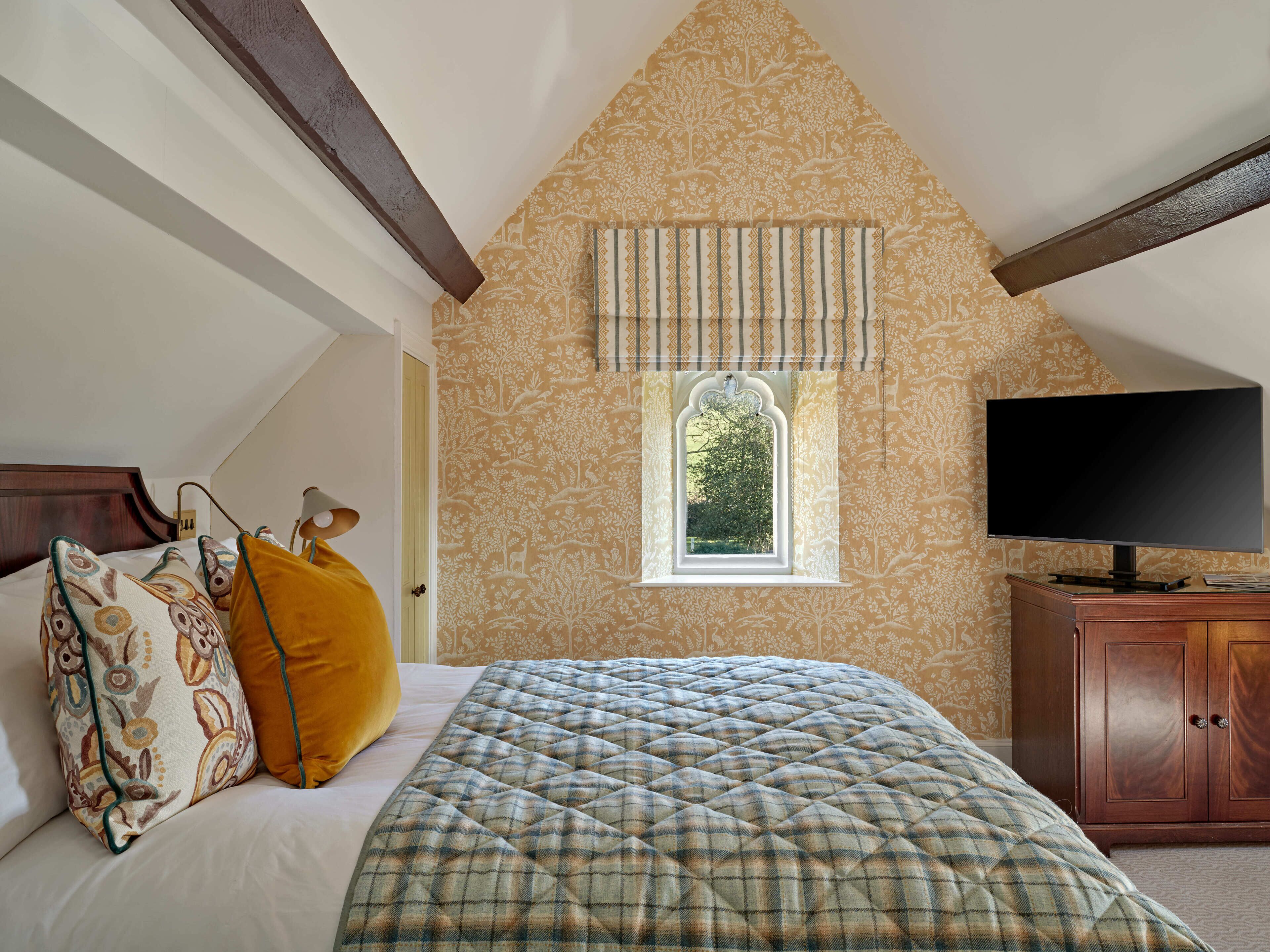 A bedroom with a sloped ceiling, a window with a decorative valance, a bed with patterned pillows, a television on a wooden cabinet, and a plaid quilt.