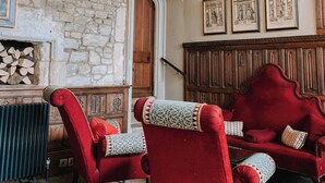 Une pièce avec un mur en pierre, une porte en bois, un lustre, des fauteuils à revêtement rouge, un tapis à motifs et des œuvres d’art encadrées accrochées au mur.