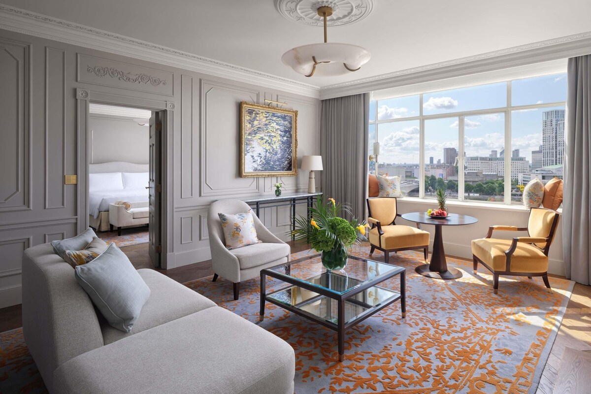 Quarto de hotel com cama, duas poltronas, mesa de centro e janela grande com vista para a cidade.
