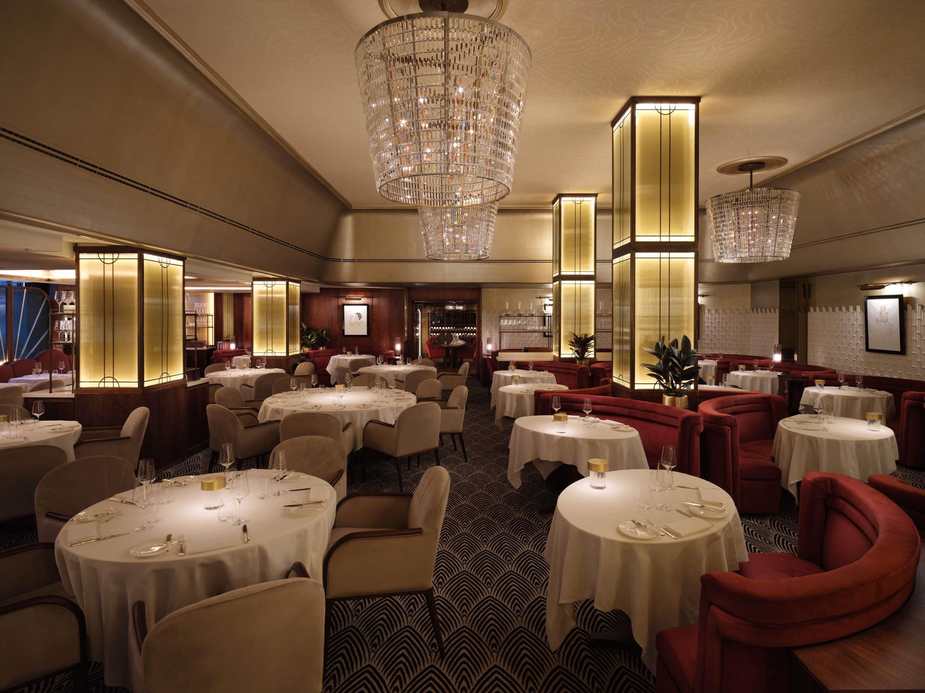 A spacious dining area with multiple round tables, elegant chandeliers, and a patterned carpet.