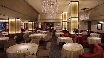 A spacious dining area with multiple round tables, elegant chandeliers, and a patterned carpet.