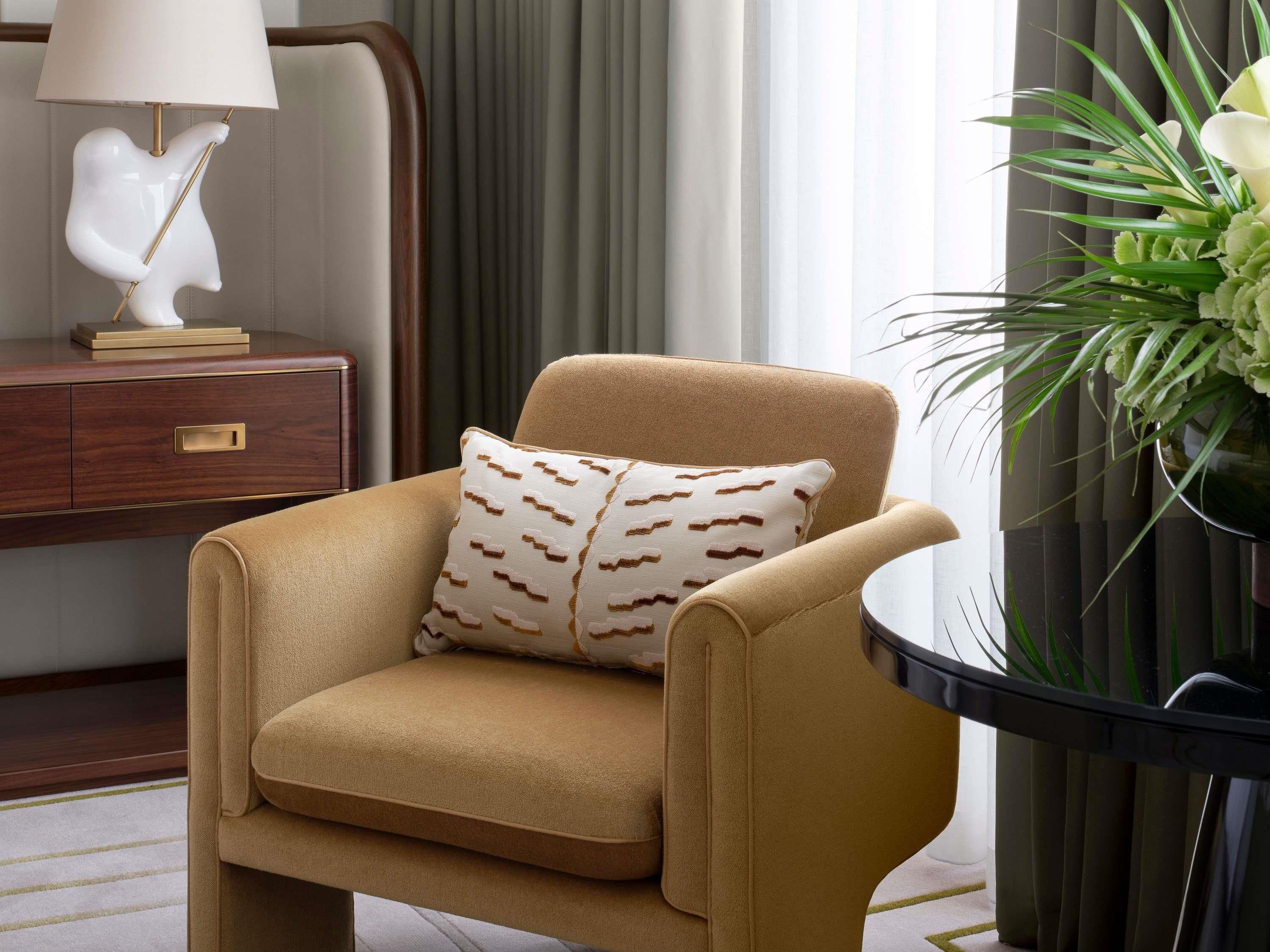 A beige armchair with a patterned pillow, a side table with a lamp, and a potted plant.
