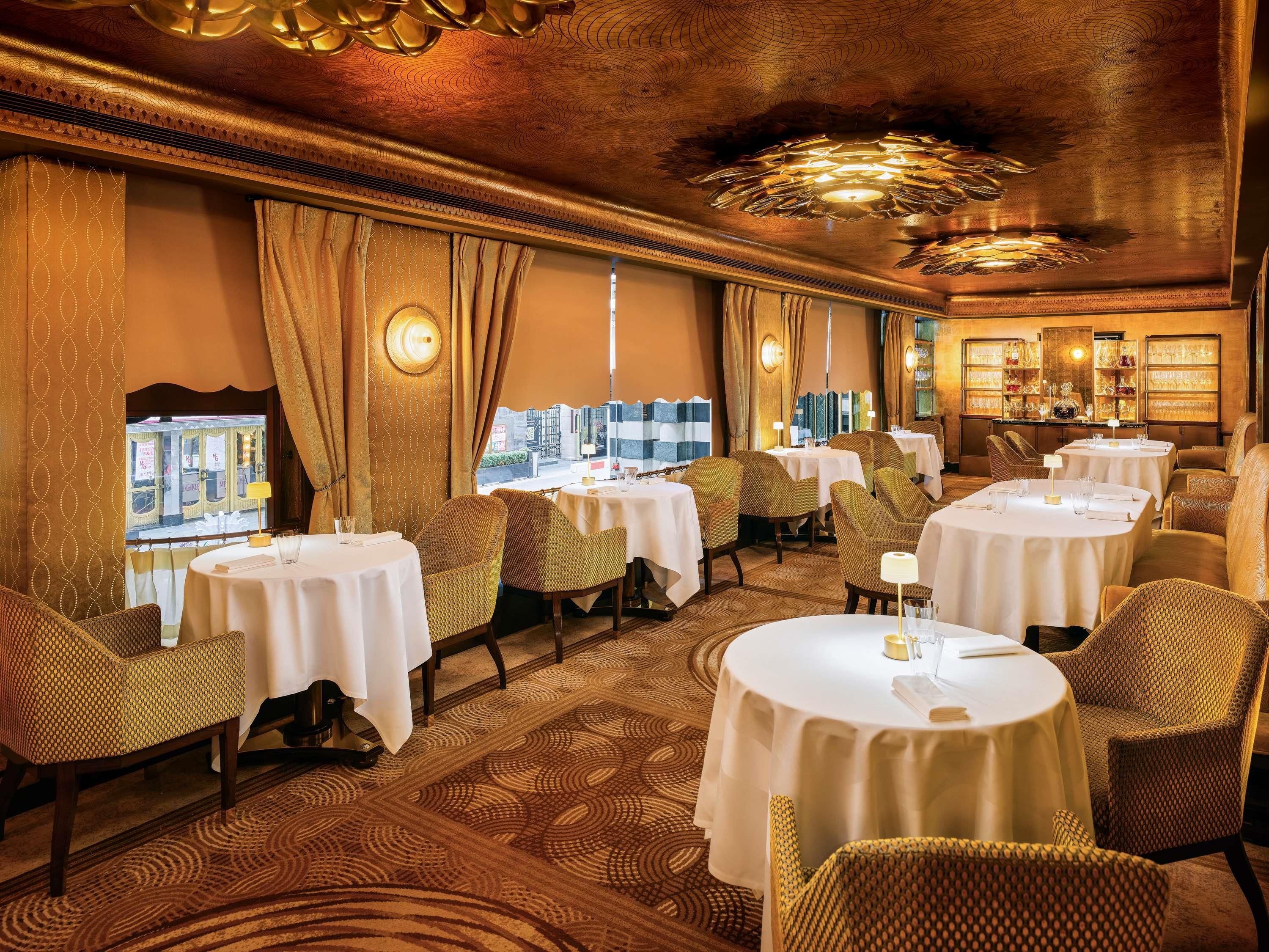 Un comedor de hotel con mesas redondas, sillas de mimbre y vistas a la ciudad a través de amplios ventanales.