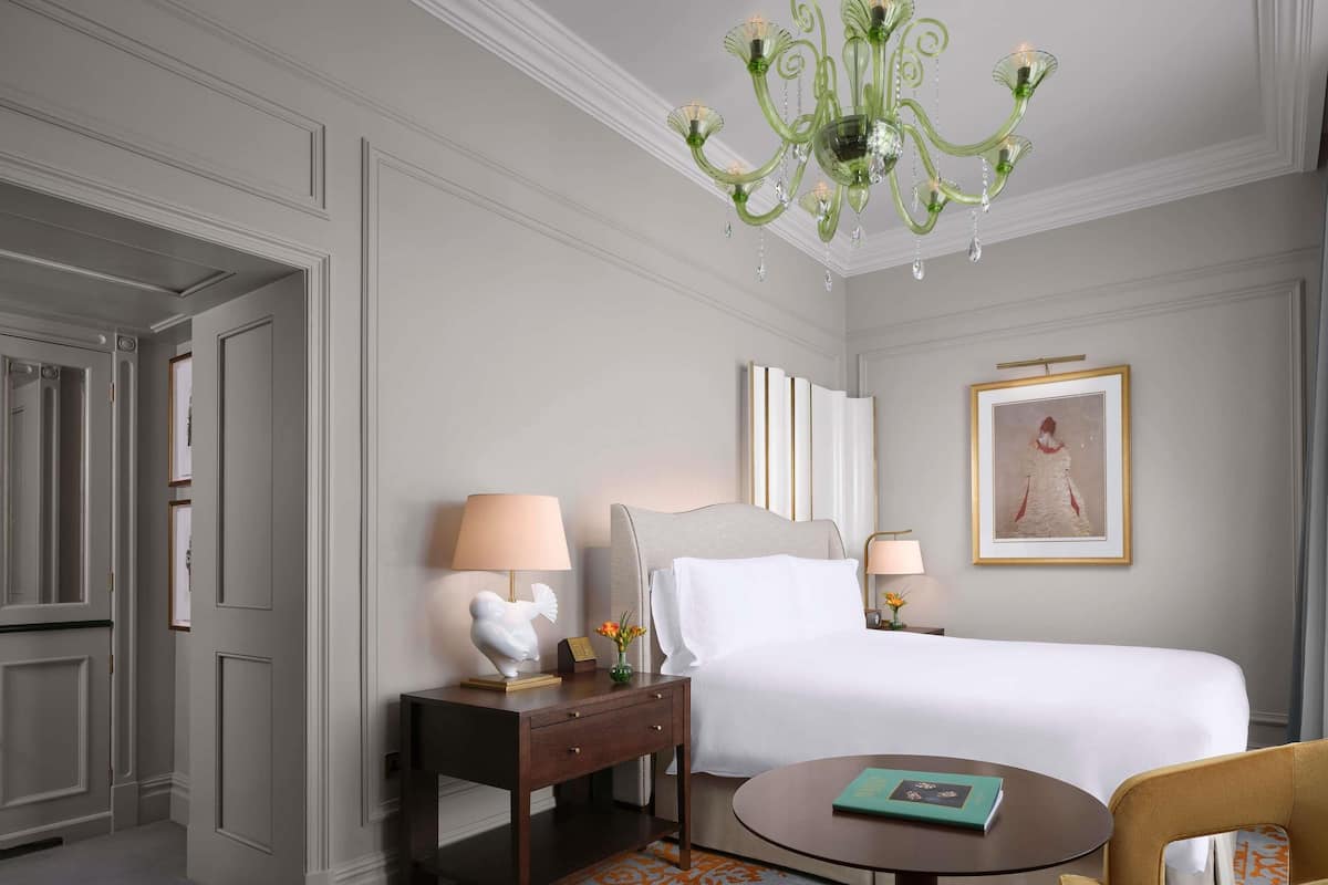 Habitación de hotel con cama, mesitas de noche, una silla, una mesa redonda, una araña y un cuadro en la pared.