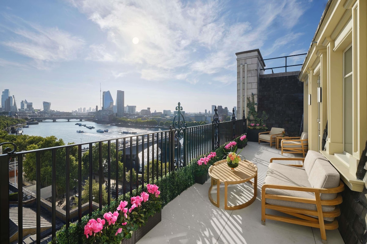 A balcony with a view of a river, city skyline, and a bridge. There are outdoor seating arrangements and a railing.