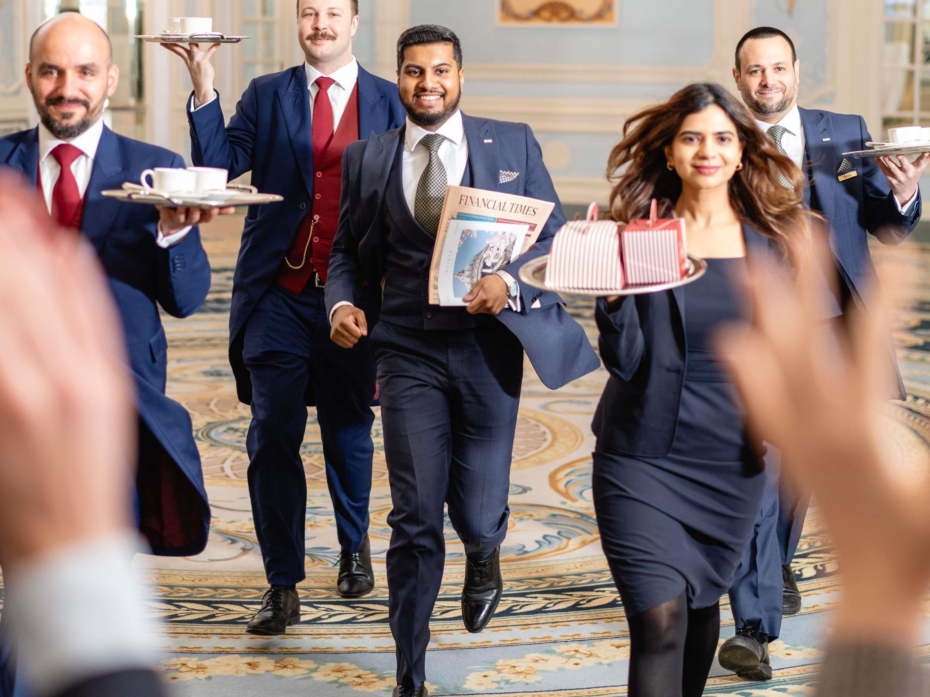 A group of formally dressed individuals carrying trays and a newspaper, walking on a patterned carpet in a room with ornate decor.
