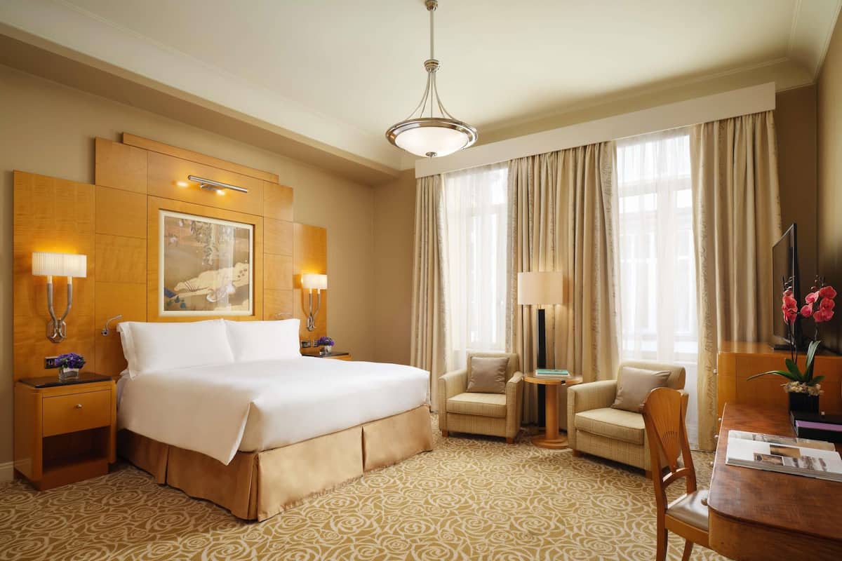 Habitación de hotel con una cama grande, dos mesitas de noche con lámparas, una pequeña mesa redonda con una silla, un televisor y un ventanal con cortinas.