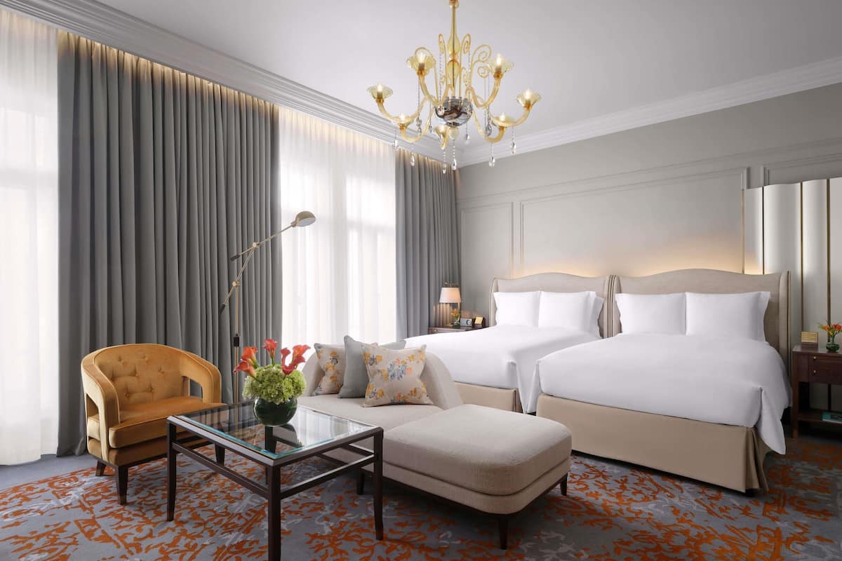 Habitación de hotel con una cama grande, una cama más pequeña, una mesa de centro de vidrio, una silla y un candelabro.