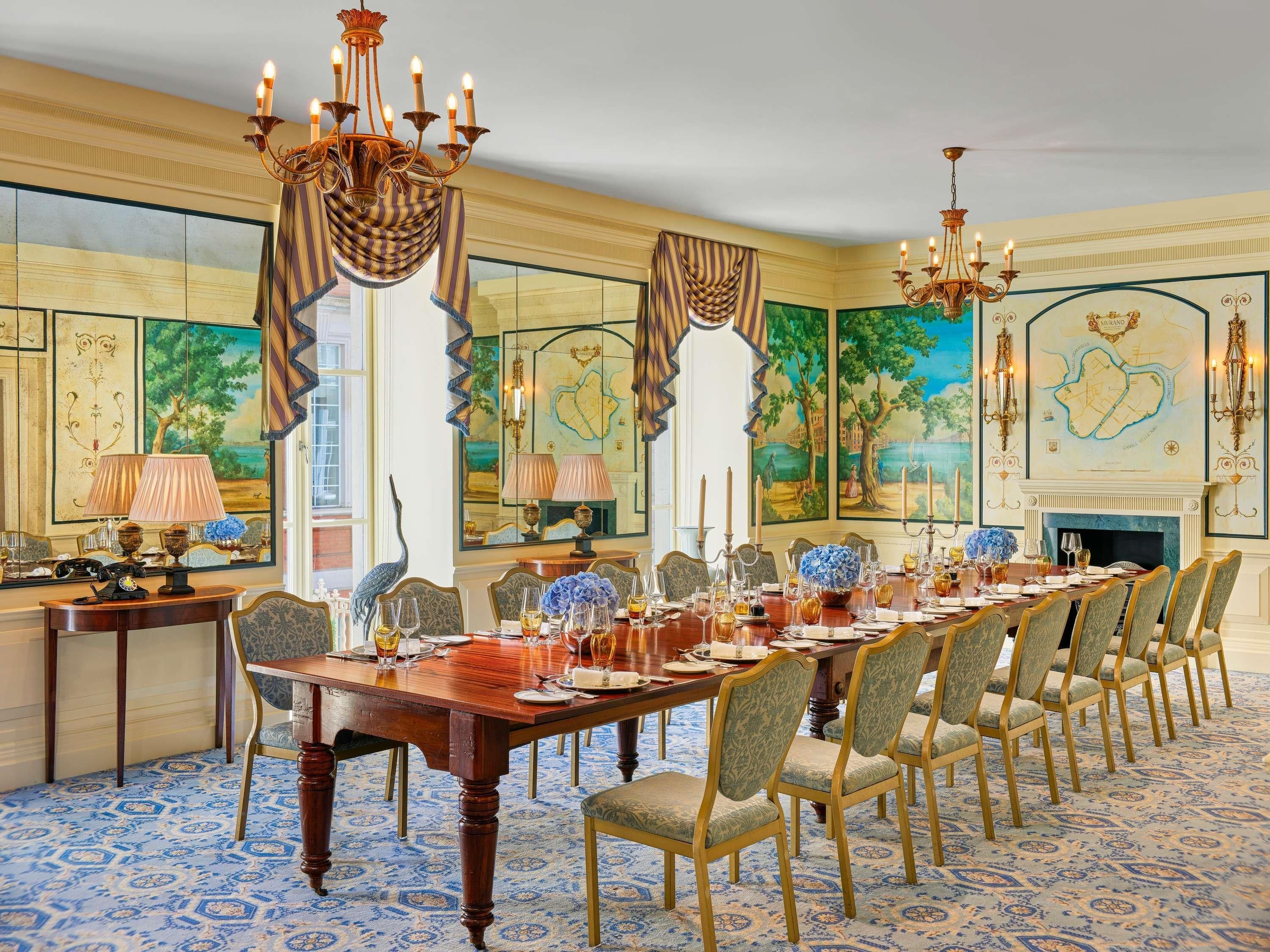 A formal dining room with a long table set for a meal, surrounded by chairs, and adorned with a chandelier and wall paintings.
