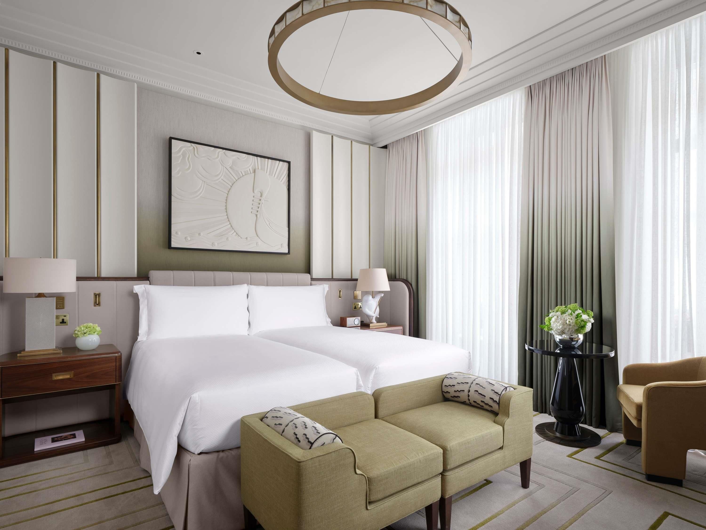 A hotel room with a double bed, two bedside tables, a sofa, a chair, a large abstract painting, and a view of the outdoors through sheer curtains.