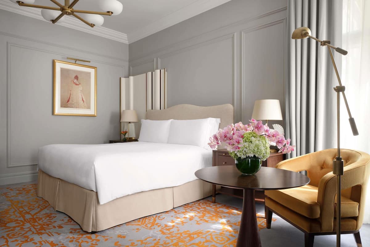 Habitación de hotel con una cama grande, una mesita de noche con lámpara, una silla, una pequeña mesa con un jarrón de flores y un aplique de techo.