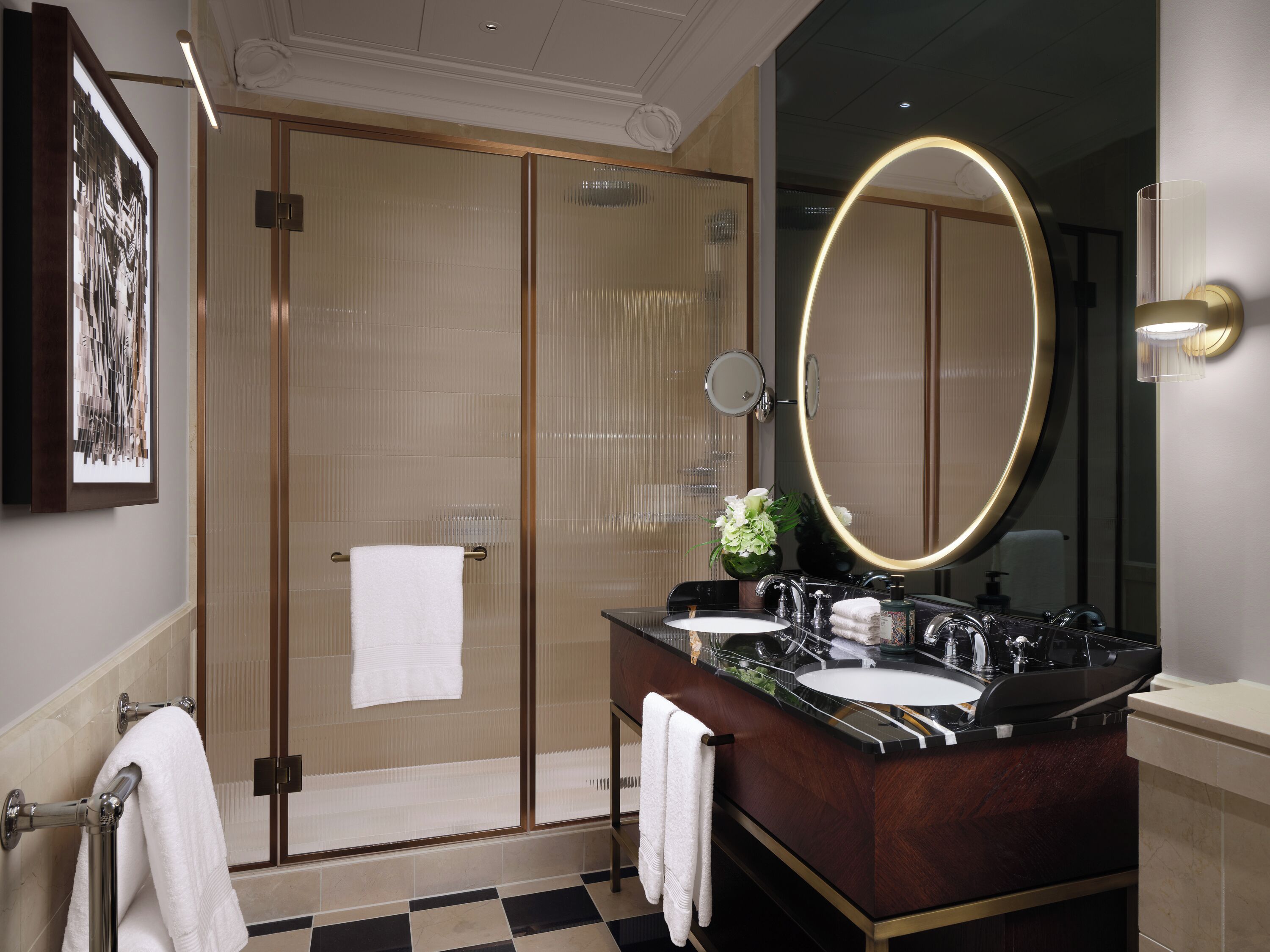 A modern bathroom with a glass-enclosed shower, a large mirror with a gold frame, and a dark marble countertop with two sinks.