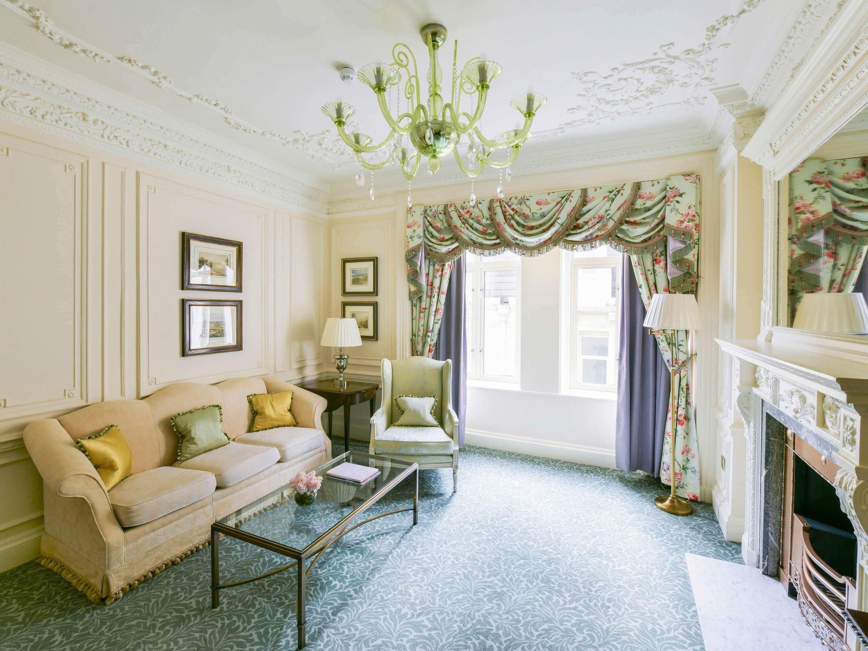 A room with a sofa, armchair, glass coffee table, fireplace, and floral curtains.