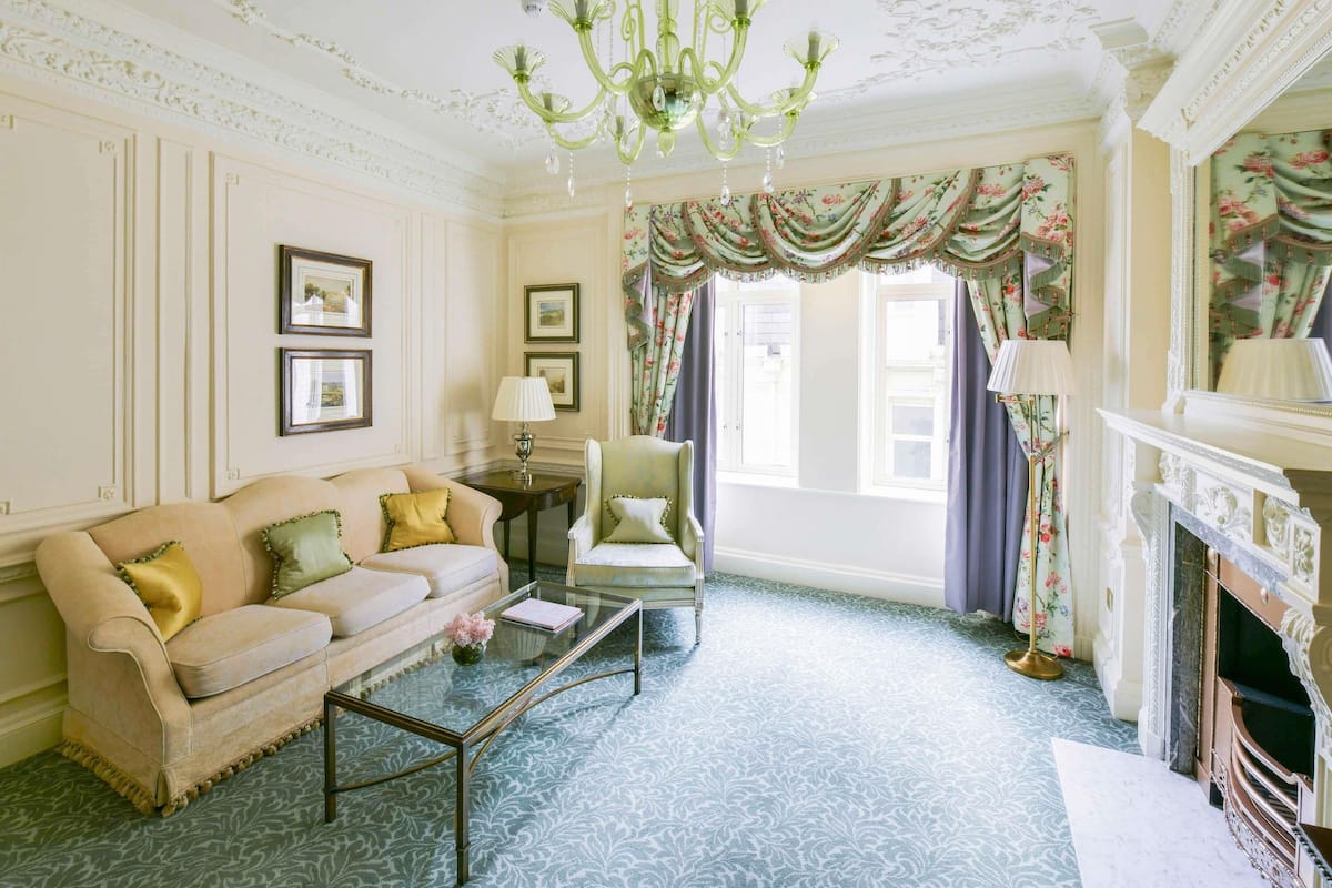 A room with a sofa, armchair, glass coffee table, fireplace, and floral curtains.