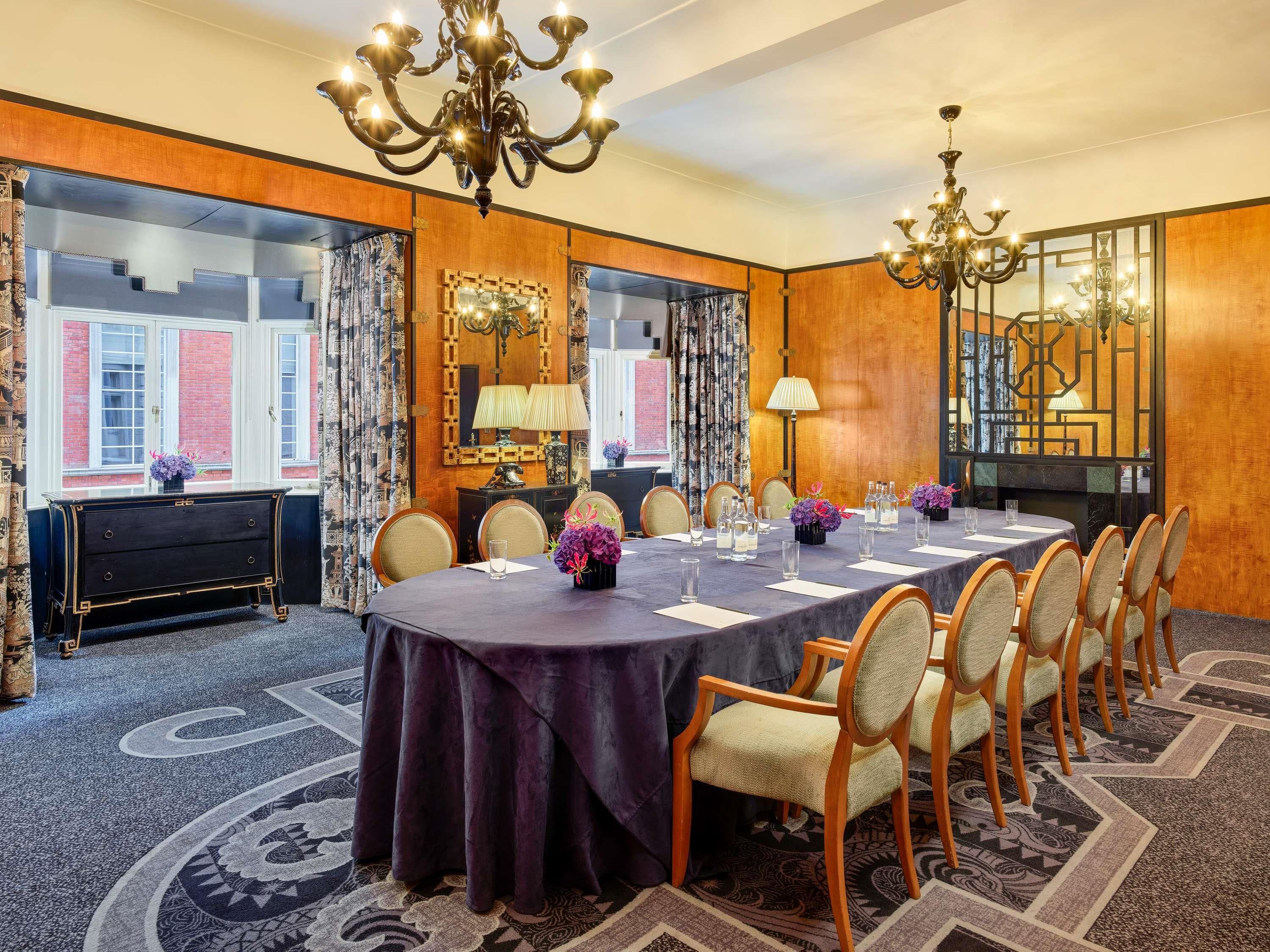 A conference room with a large round table, chairs, a chandelier, and a view of a brick building.