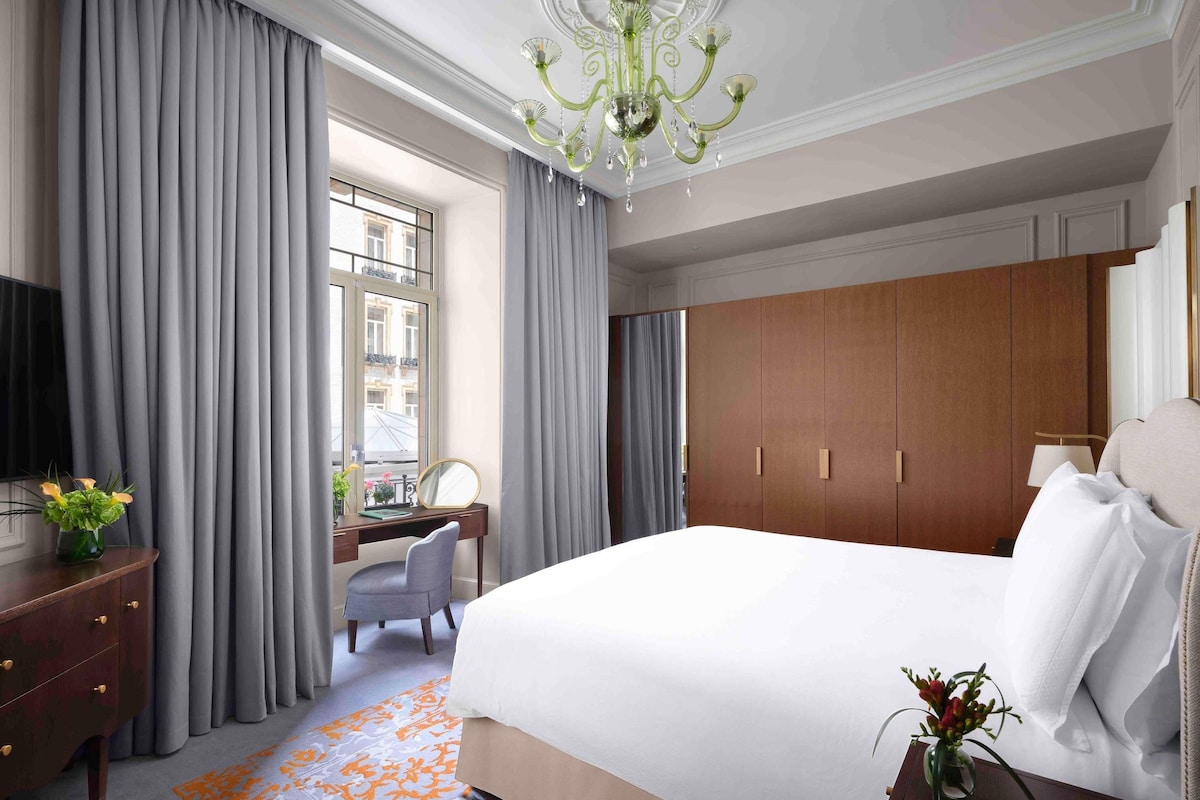 Quarto de hotel com uma cama grande, uma mesa com cadeira, televisão e janela com cortinas.