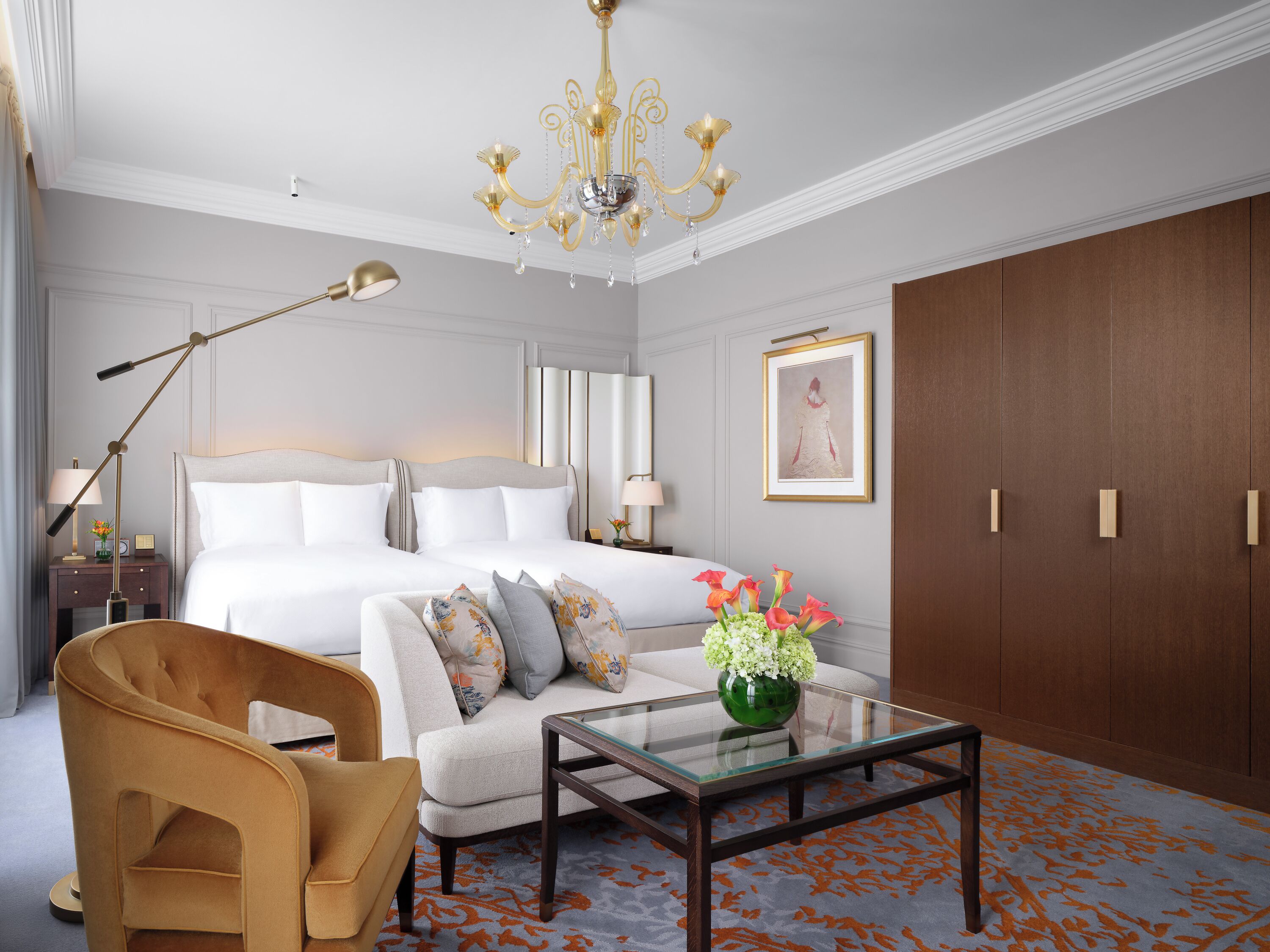 A hotel room with a large bed, a sitting area with a sofa and armchair, a glass coffee table, a chandelier, and a wardrobe.