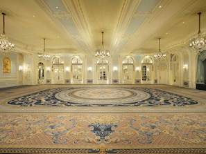 A grand hall with ornate chandeliers, a patterned carpet, and a large, circular floor design.