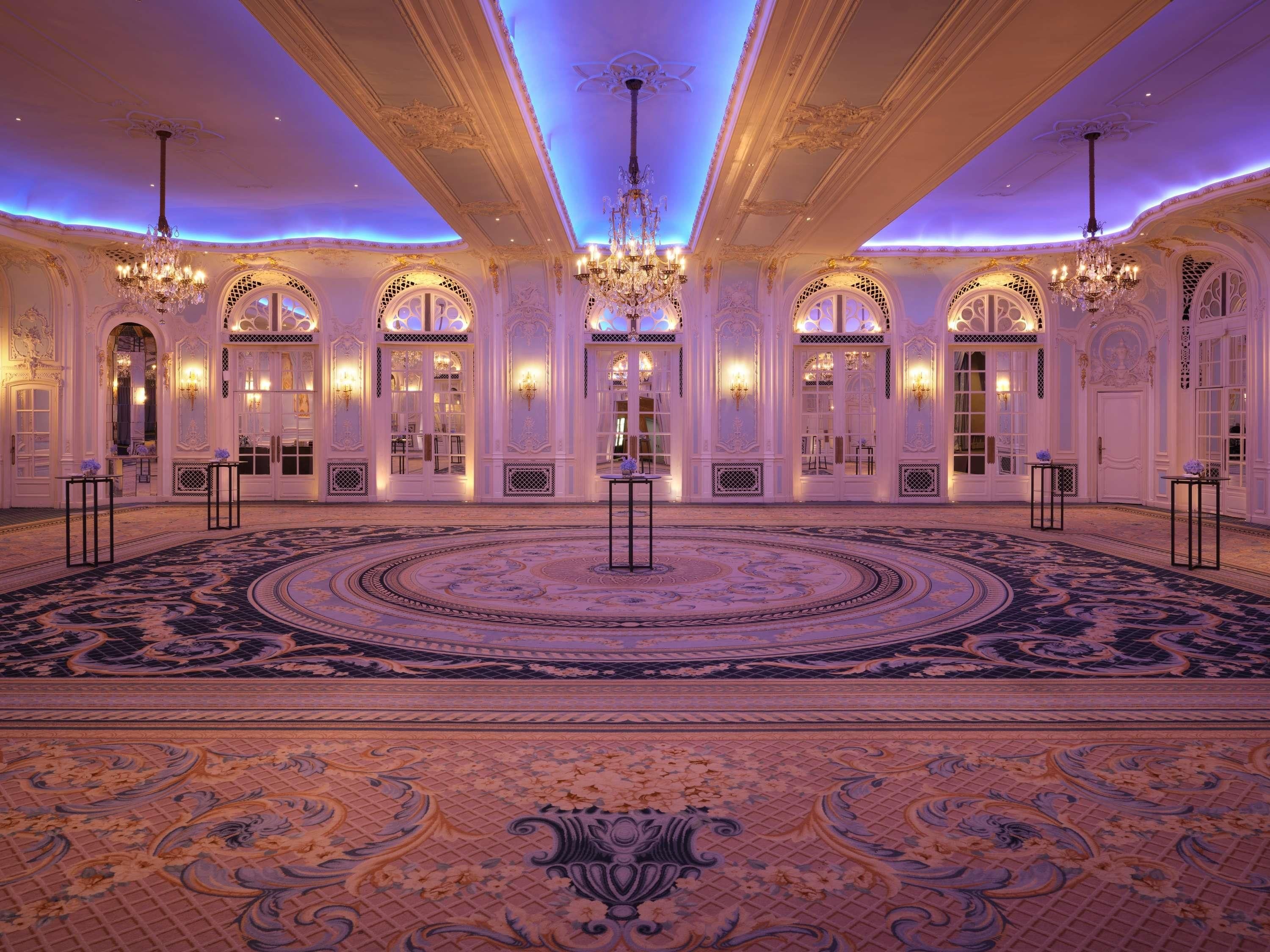 A grand ballroom with ornate chandeliers, large windows, and a patterned carpet.