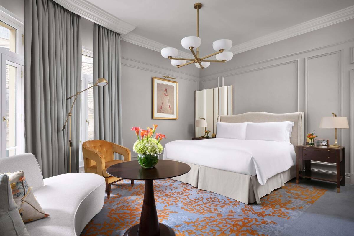 Una habitación de hotel con cama, una silla, una mesa, una lámpara, un jarrón con flores y un cuadro en la pared.