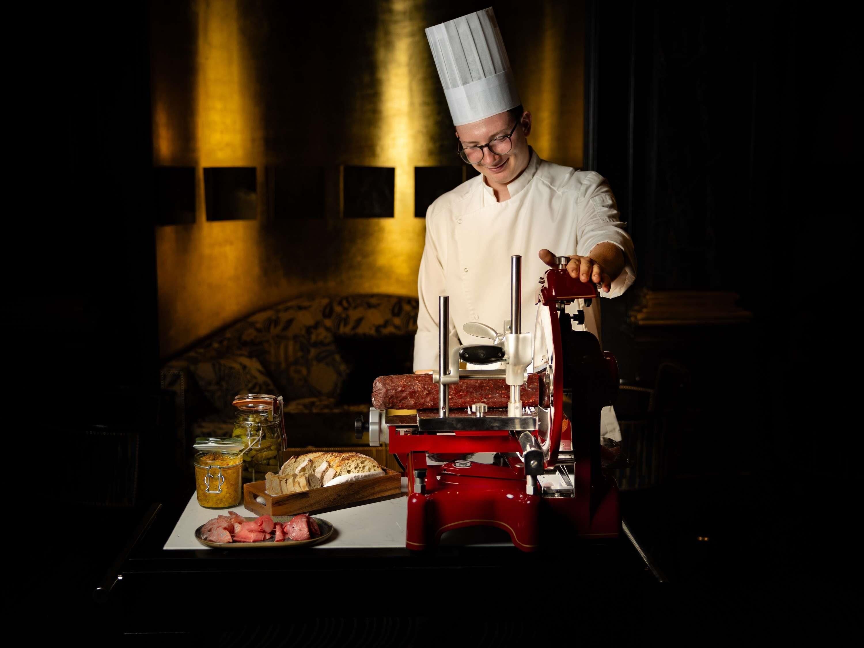 Um chef com uniforme branco e chapéu de chef alto, em frente a um processador de alimentos vermelho, com diversos ingredientes sobre o balcão.