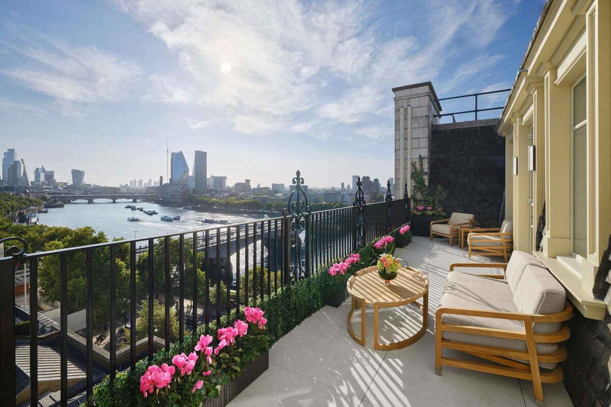 A balcony with a view of a river, city skyline, and a bridge. There are outdoor seating arrangements and a railing.