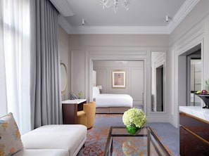 A hotel room with a bed, a sitting area with a chair and a small table, a mirror, and a vase with flowers. - The Savoy (London)