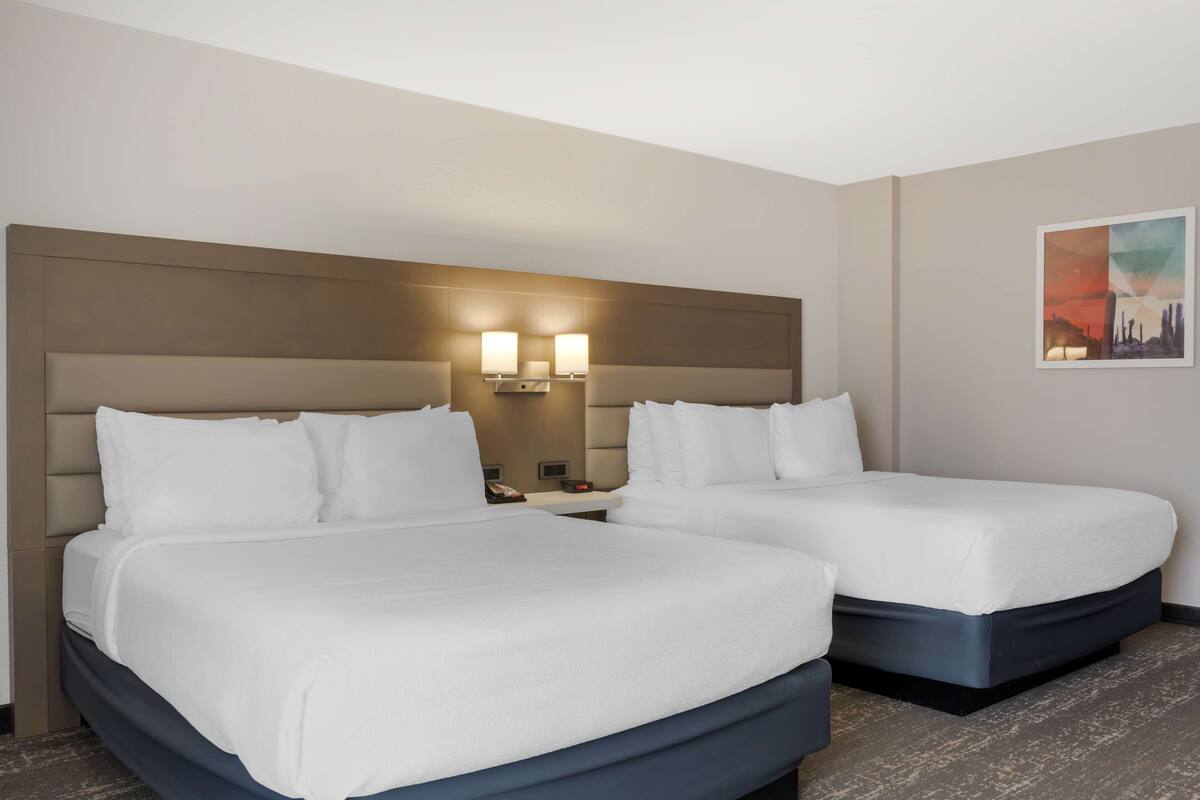 A hotel room with two beds, a headboard, wall-mounted lights, a nightstand, and a painting.