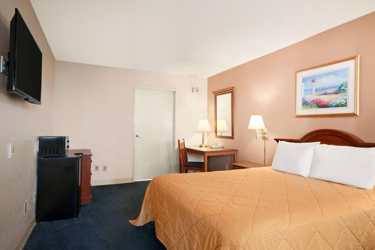 A hotel room with a bed, a desk with a chair, a television, a refrigerator, and a painting on the wall.