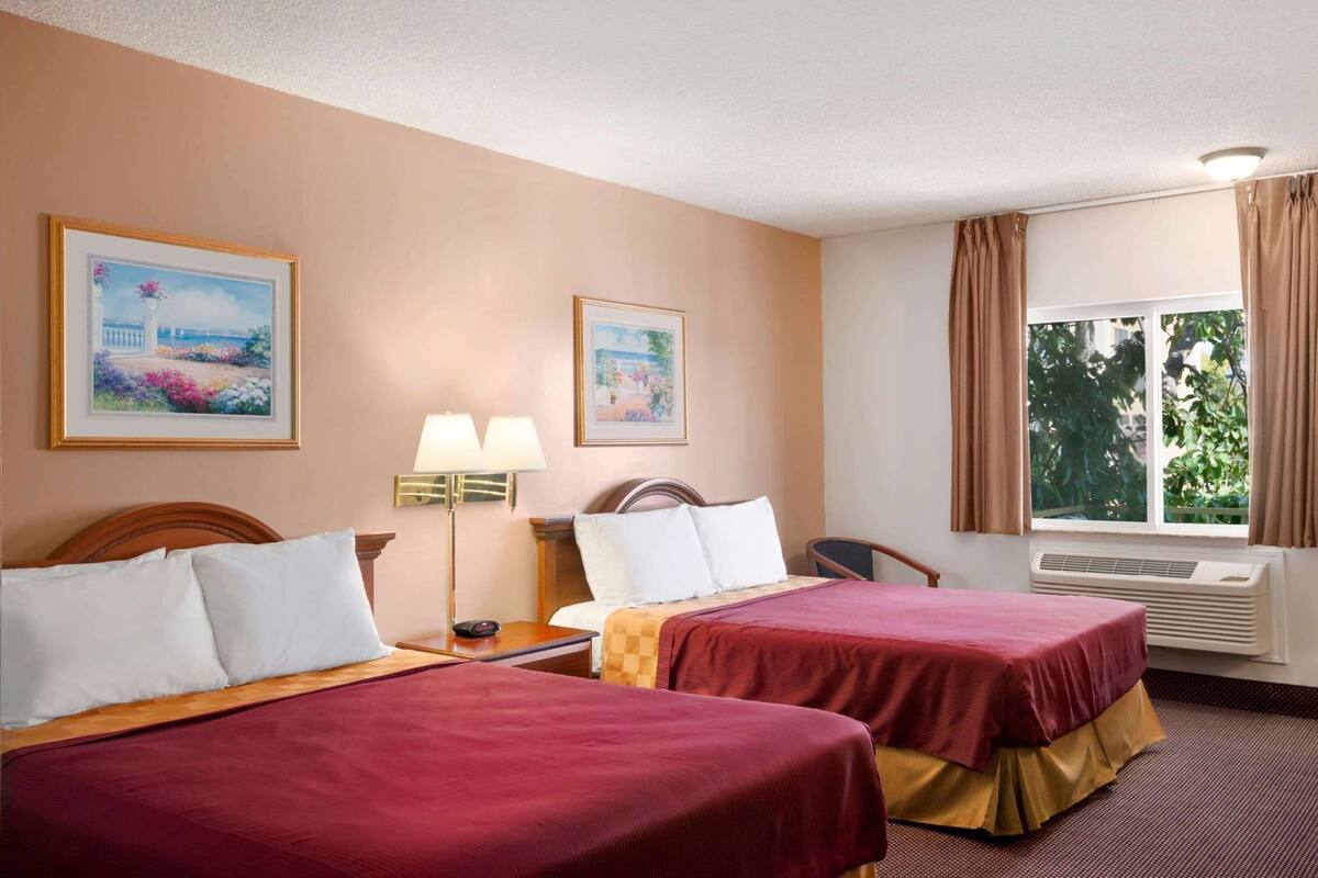 A hotel room with two beds, a nightstand, a lamp, two framed pictures, a window with curtains, and an air conditioning unit.