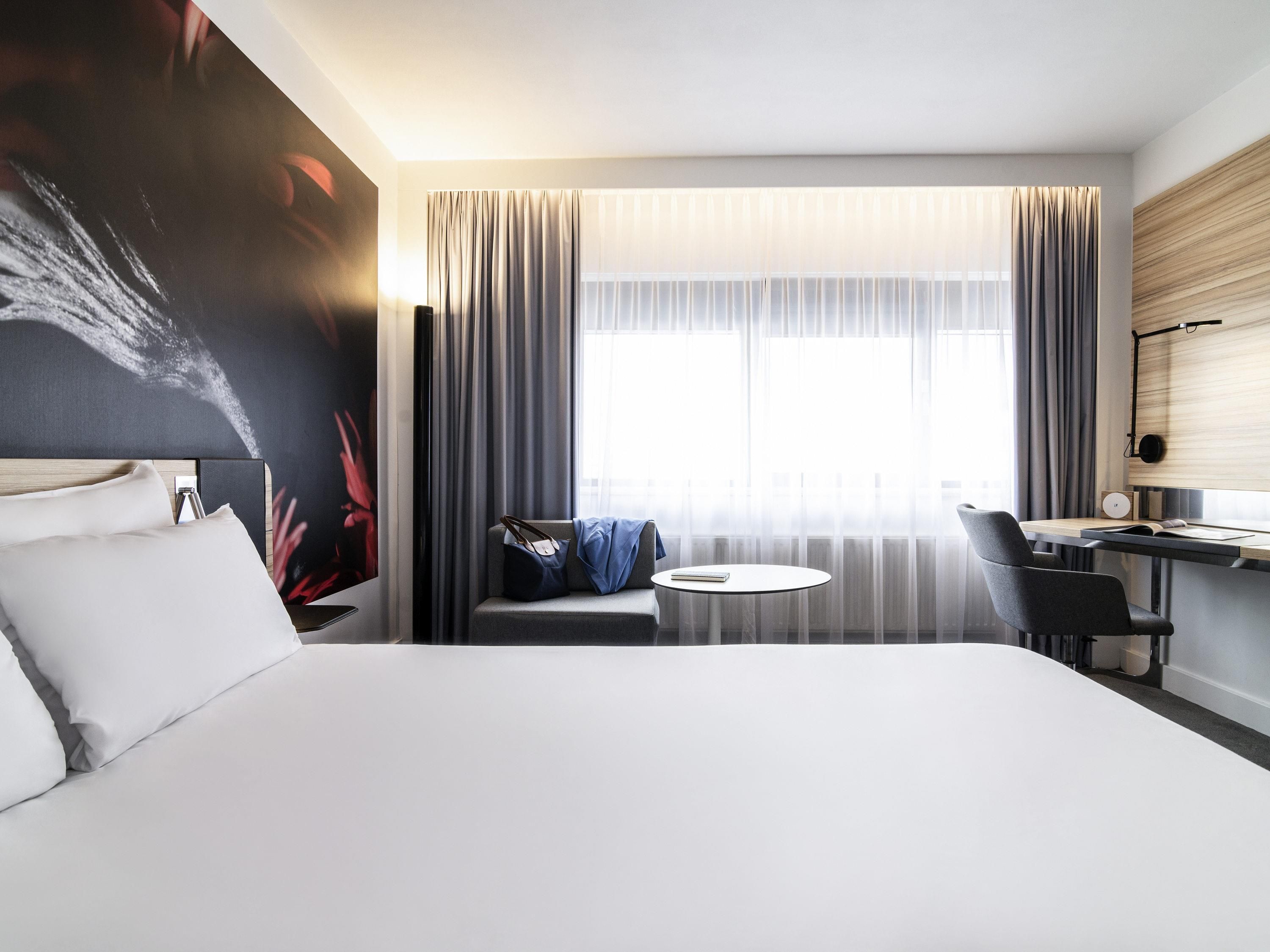 A modern hotel room with a large bed, a desk with a chair, a small round table, and a large window with curtains.