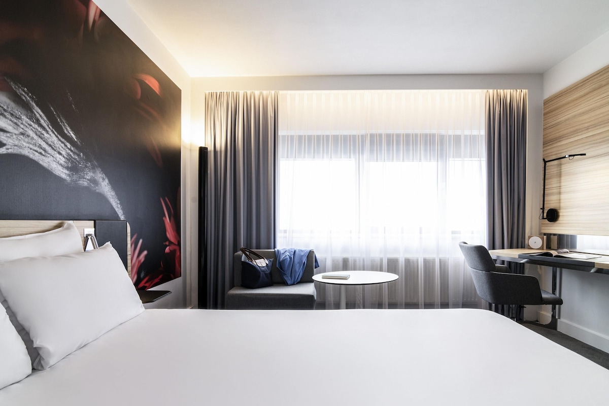 Habitación de hotel moderna con una cama grande, un escritorio con silla, una mesita redonda y un amplio ventanal con cortinas.