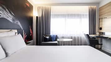 A modern hotel room with a large bed, a desk with a chair, a small round table, and a large window with curtains.