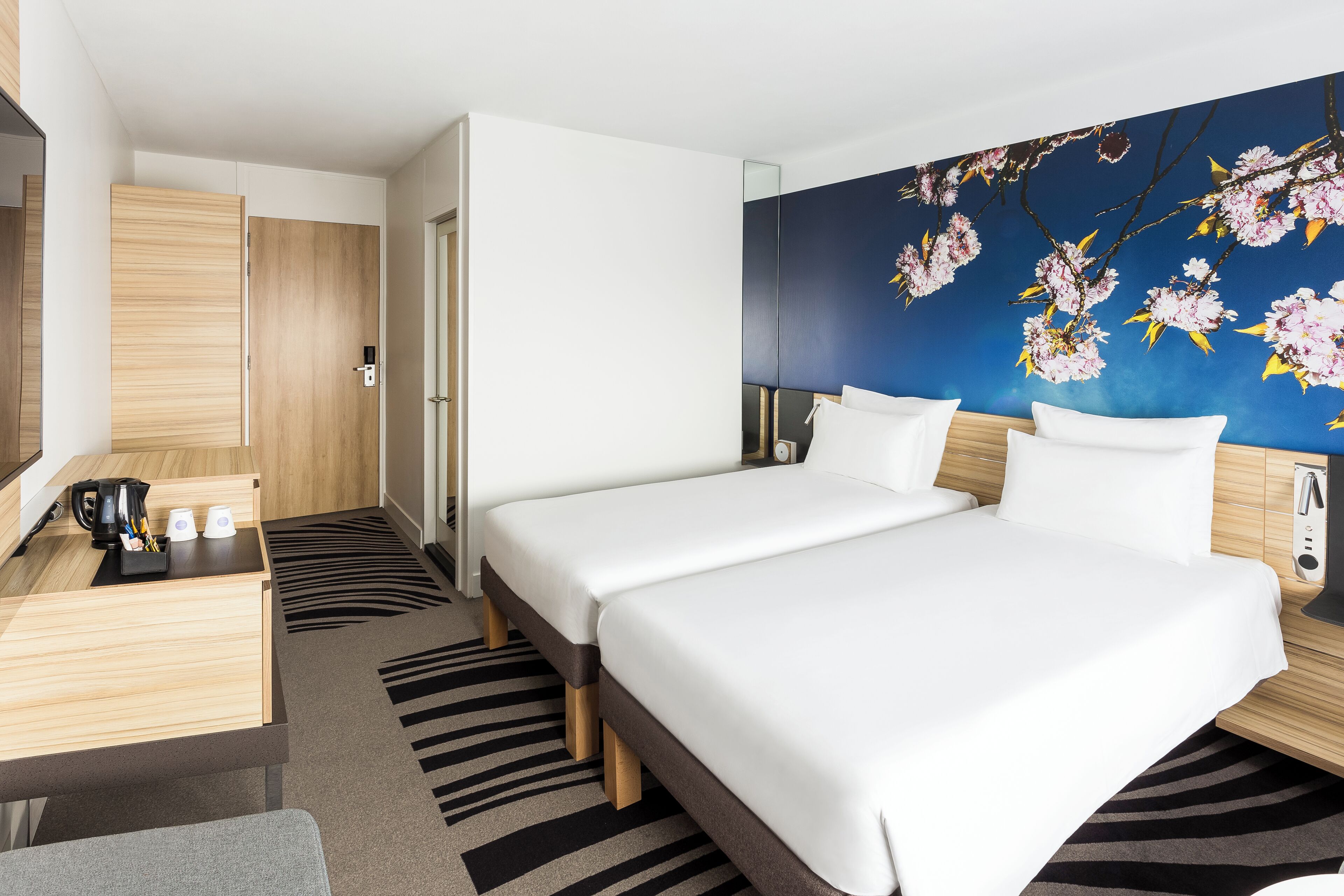 A hotel room with two beds, a desk with a coffee maker, a mirror, and a wall with a floral mural.