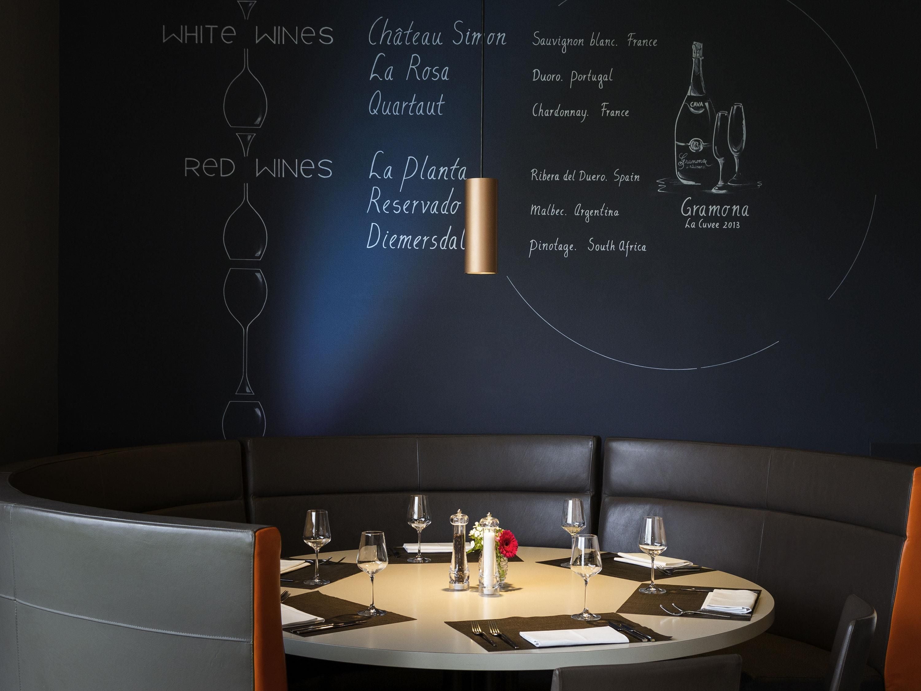Interior de un restaurante con un menú en una pizarra que enumera vinos blancos y tintos, una mesa redonda lista para comer y asientos en cabinas de cuero.