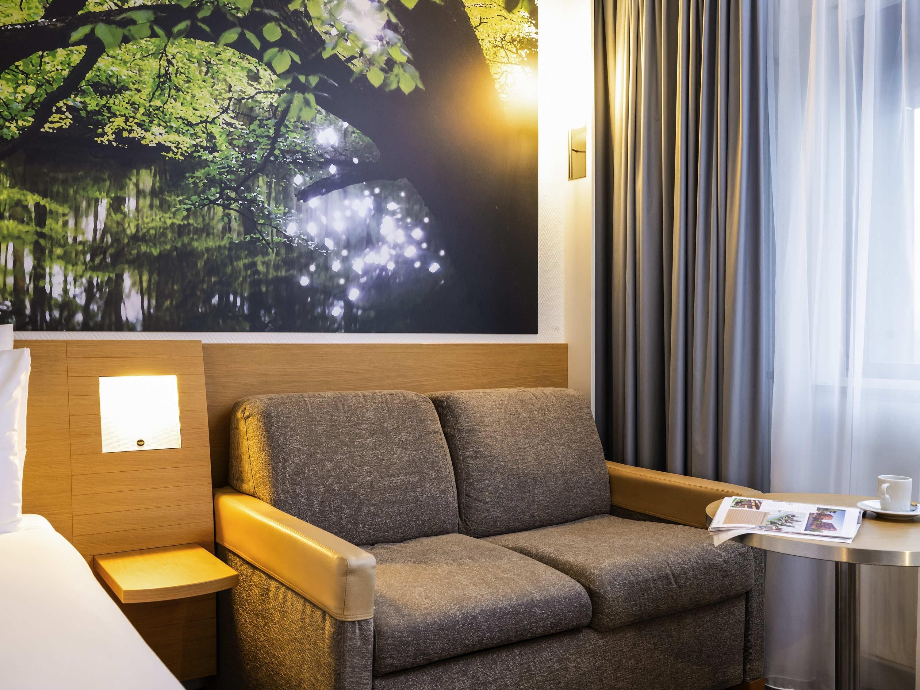 A hotel room with a large wall mural of a tree and water, a sofa, a bedside table with a lamp, and a small table with a magazine and a cup.