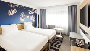 A hotel room with two beds, a desk, a TV, and a large window with curtains. - Novotel Amsterdam City (Amsterdam)