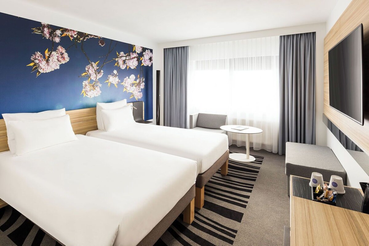 Ein Hotelzimmer mit zwei Betten, einem Schreibtisch, einem Fernseher und einem großen Fenster mit Vorhängen.