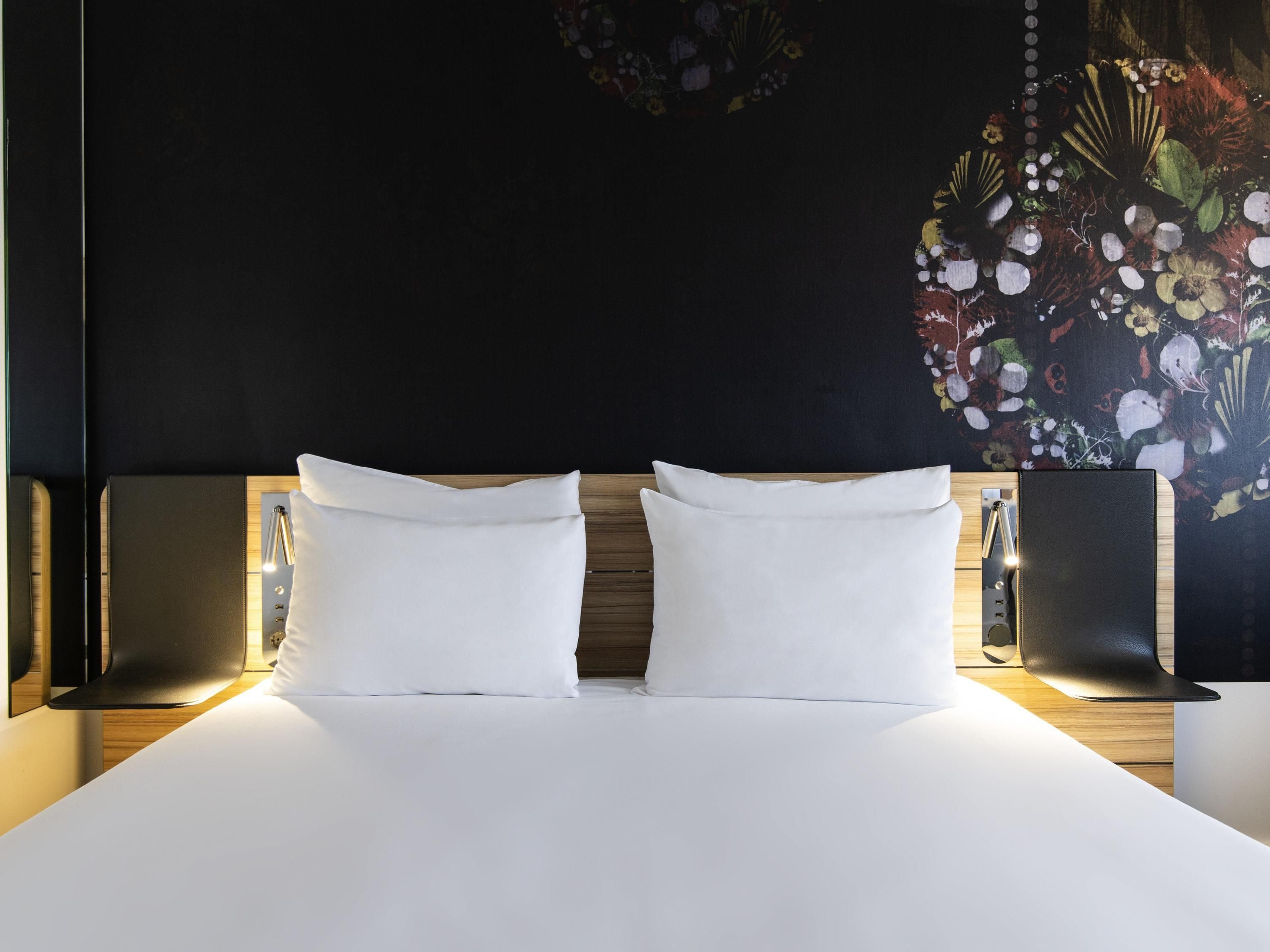 Ein Hotelzimmer mit einem großen Bett, zwei weißen Kissen, einer dunklen Wand mit floralem Wandbild und zwei schwarzen Lautsprechern auf beiden Seiten des Bettes.