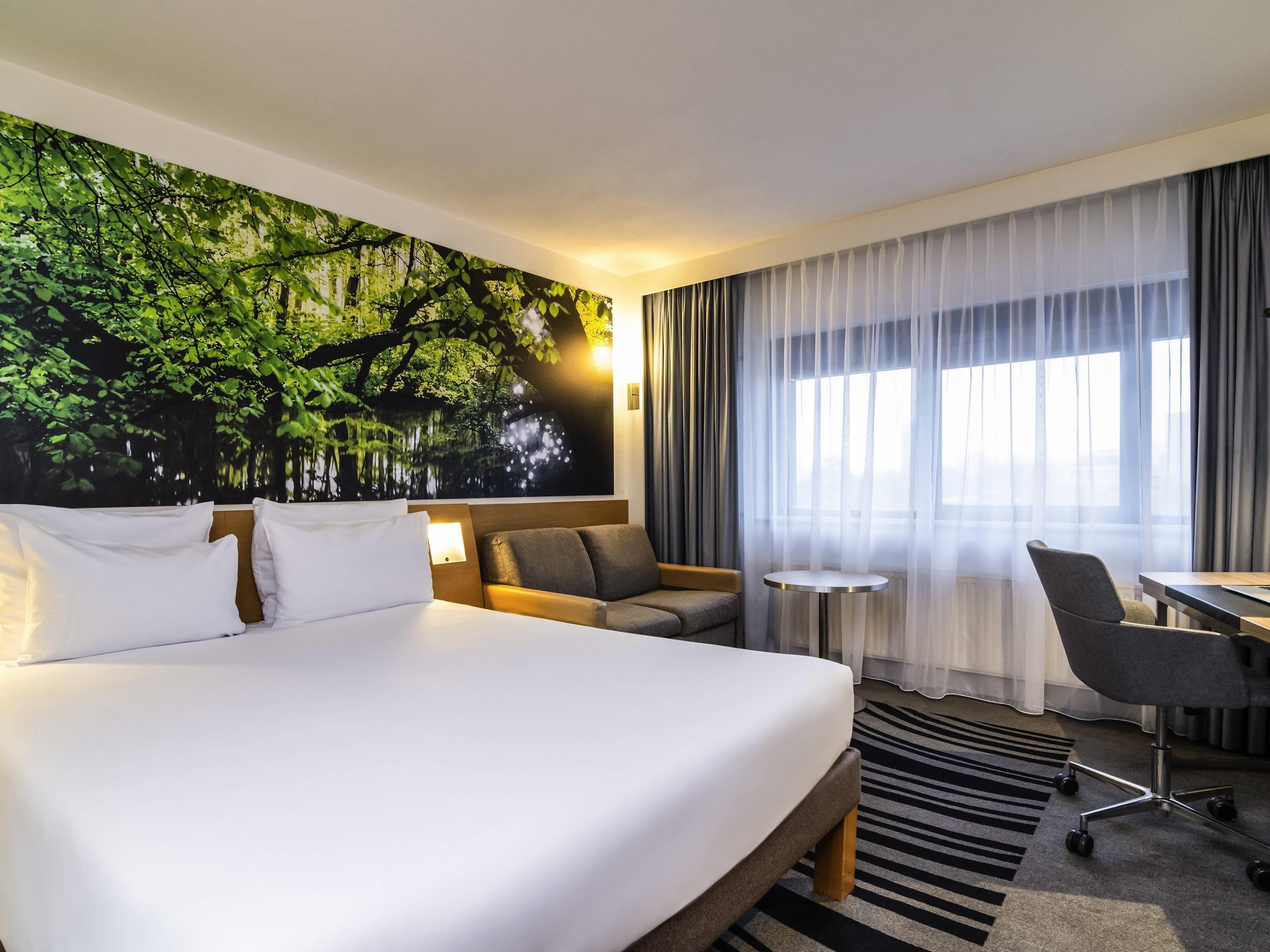 A hotel room with a large bed, a sofa, a desk, and a chair. There is a window with sheer curtains and a view of the outside.