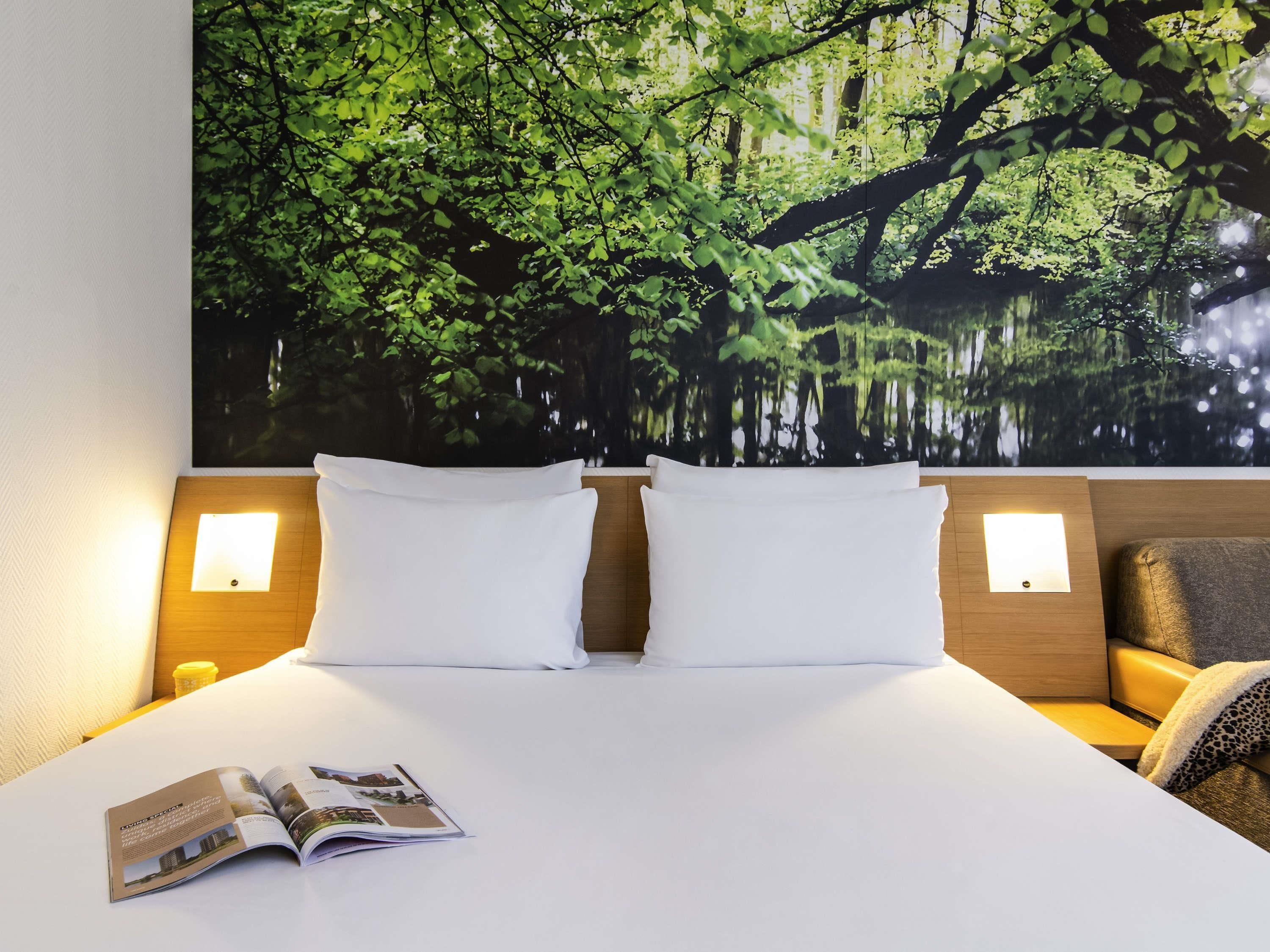 Una habitación de hotel con una cama grande, dos almohadas, una revista sobre la cama, dos lámparas de noche y un mural grande con motivos forestales.