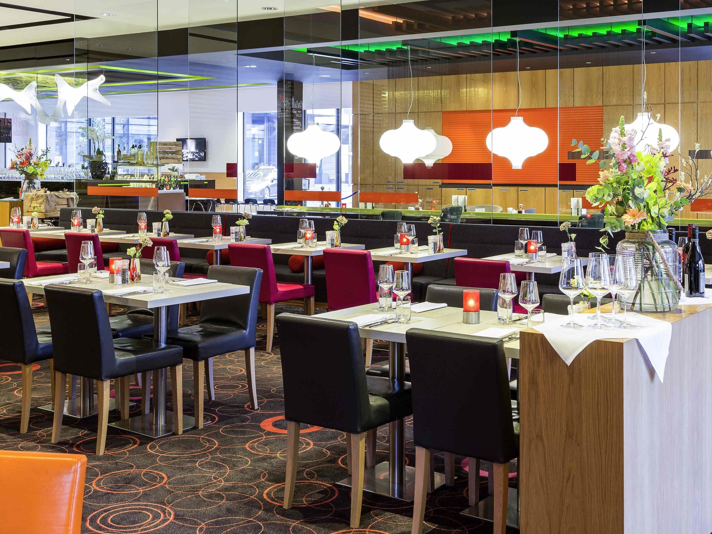 Un restaurant moderne avec des tables dressées pour le dîner, où l’on trouve un mélange de chaises rouges et noires, ainsi qu’un vase décoratif avec des fleurs posé sur la table.