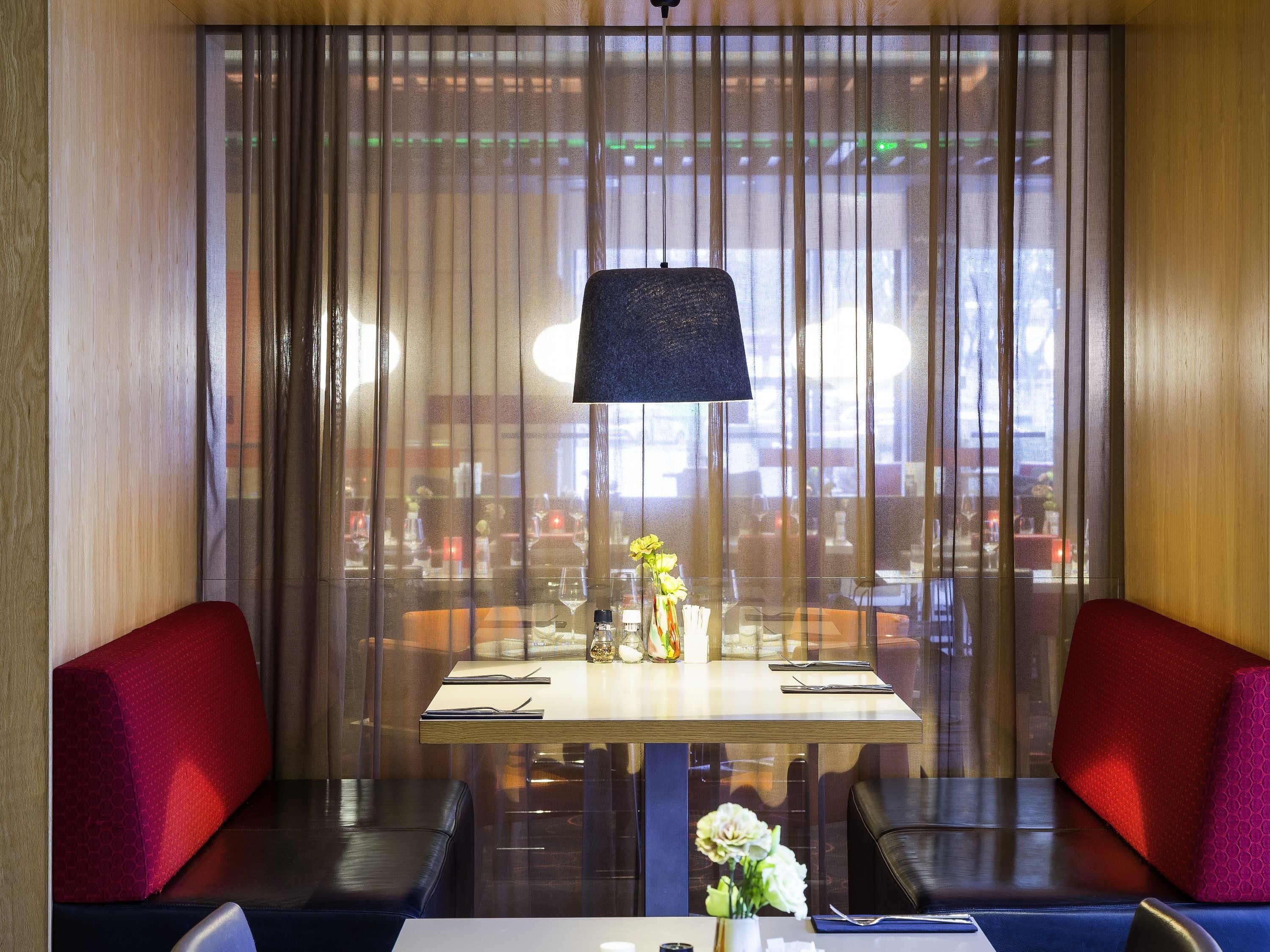 Un restaurante con una mesa para dos, un jarrón de flores y una vista a través de una ventana con persianas verticales.